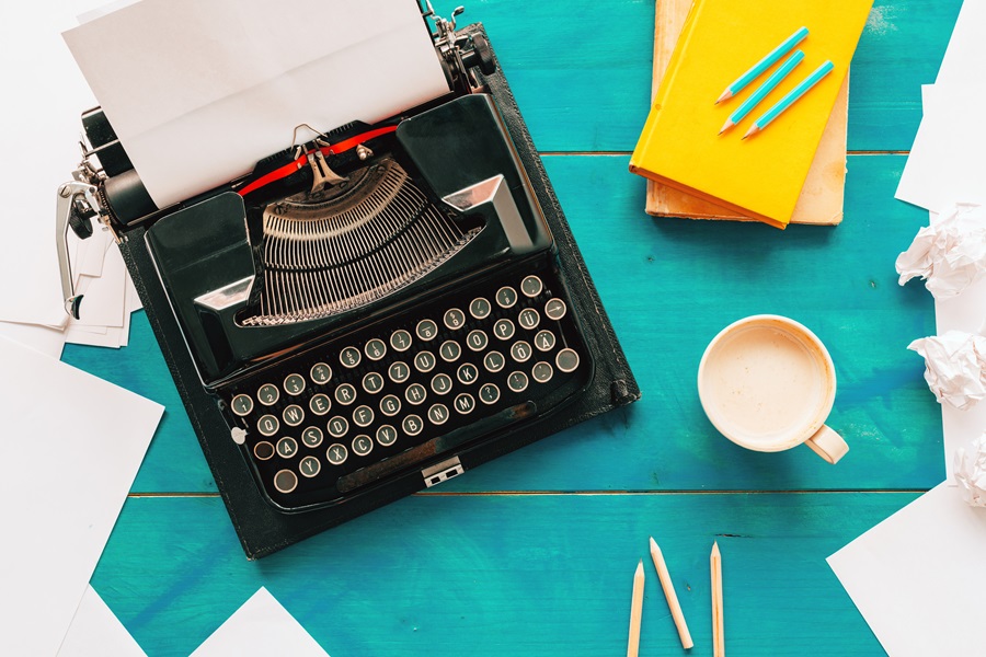 Overhead shot of a typewriter next to a coffee mug, some pencils, and a couple of notebooks.